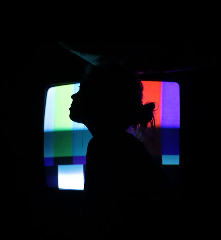 A person’s silhouette in front of a brightly lit TV screen displaying colorful test patterns in a dark room.