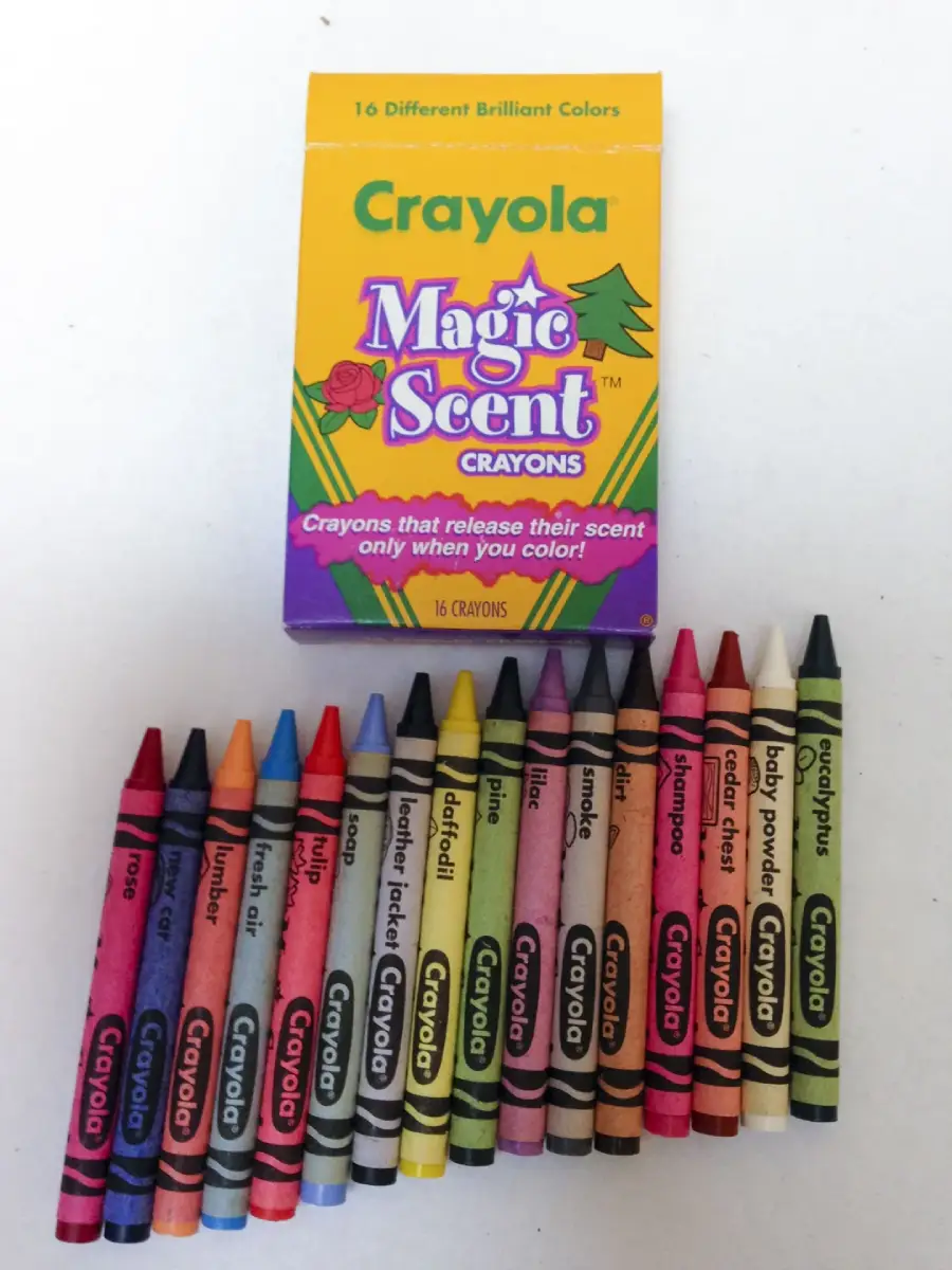 A box of Crayola Magic Scent Crayons is shown above 16 crayons arranged in a row. Each crayon is labeled with a color and scent, such as watermelon, rose, and coconut, with colorful wrappers.