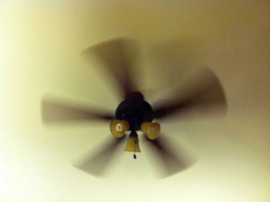 A ceiling fan with four blades is spinning, creating a motion blur effect. The ceiling is beige, and the fan has three lightbulbs and a pull chain hanging down.