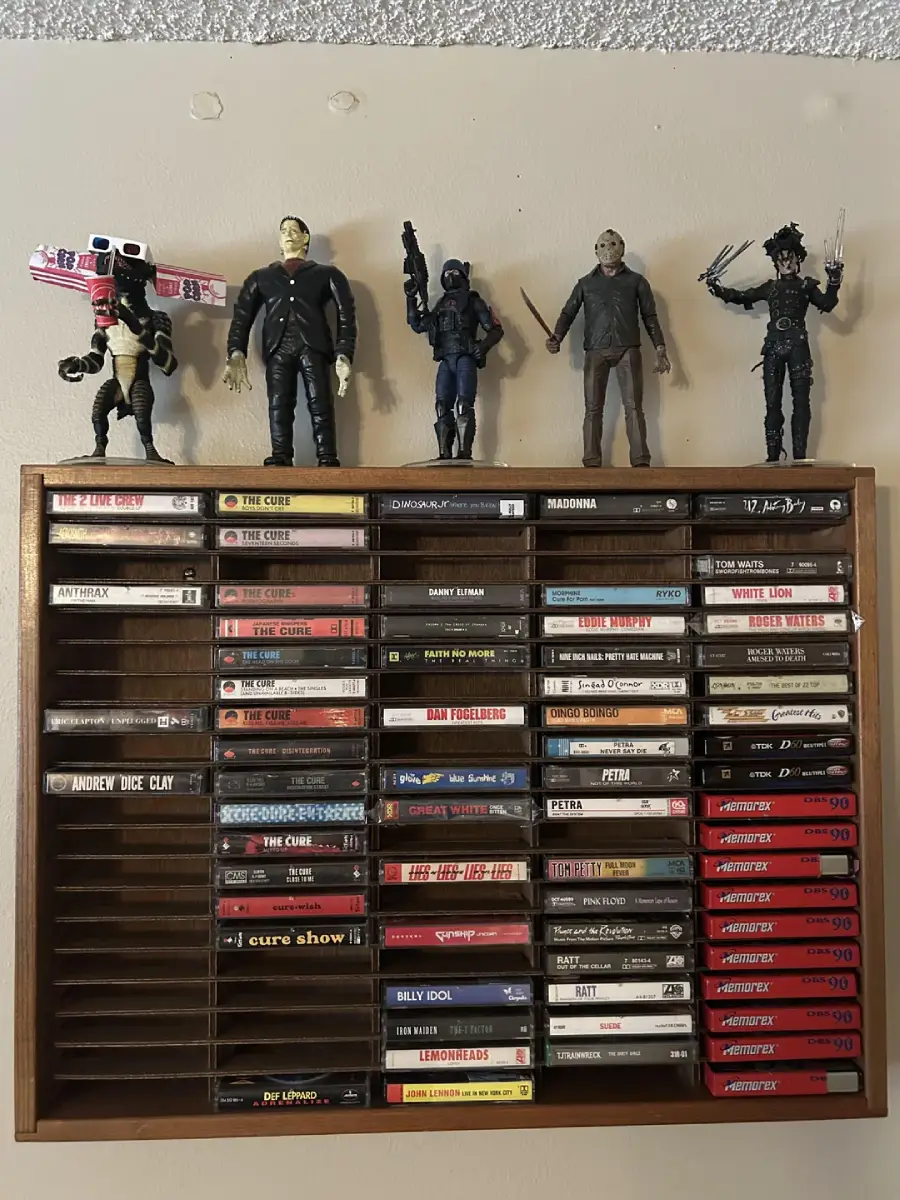 A wall-mounted wooden shelf holds rows of cassette tapes, organized neatly. On top of the shelf stand five action figures of various movie characters, posed in a line.
