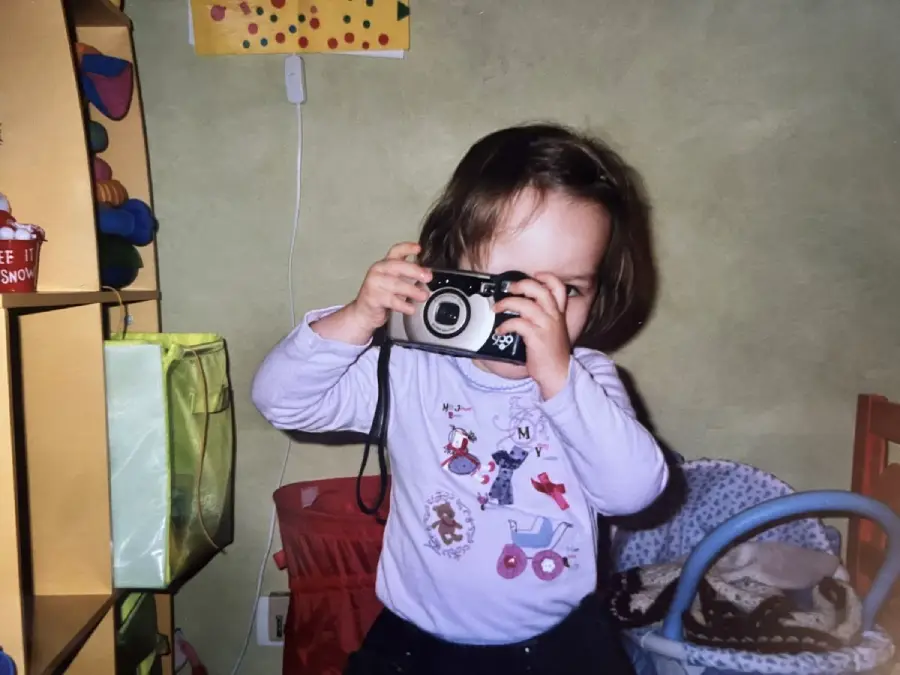A young child with brown hair wearing a light purple shirt holds a camera to their face, as if taking a picture, in a room with toys and colorful storage bins.