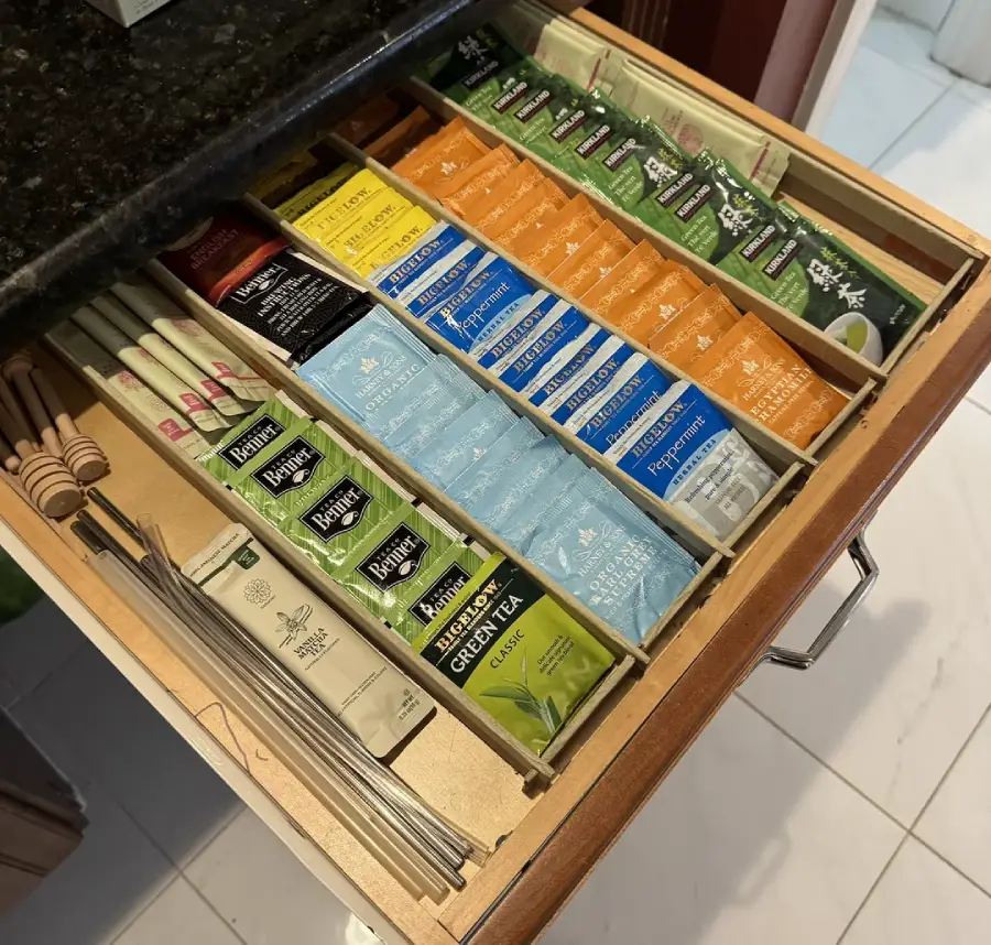 An open drawer neatly organized with assorted tea bags, individually sorted by type and brand, along with metal straws and wooden stirrers on the left side, on a light wooden divider.