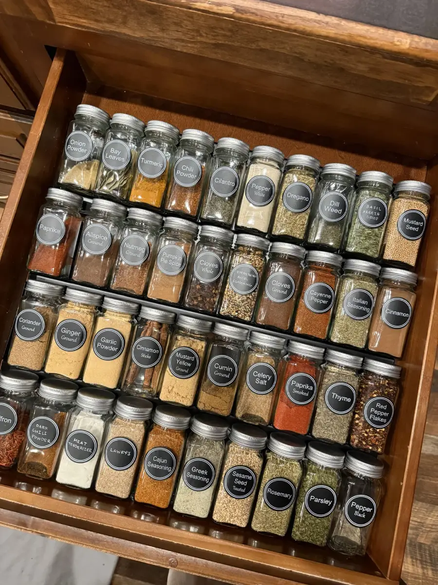 A neatly organized wooden drawer filled with labeled glass jars containing various spices and herbs, arranged in rows with round black labels on the lids.