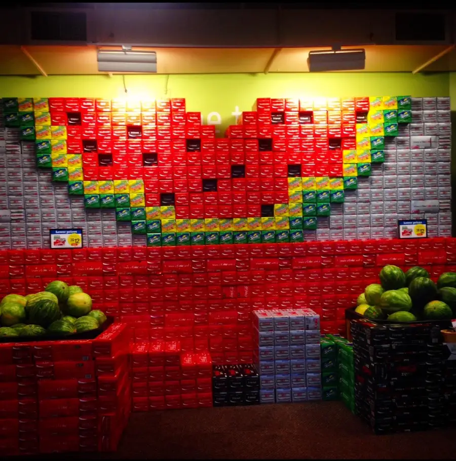 Stacks of soda boxes arranged to form a large watermelon slice on a store display, with watermelons placed in bins on either side at the bottom.