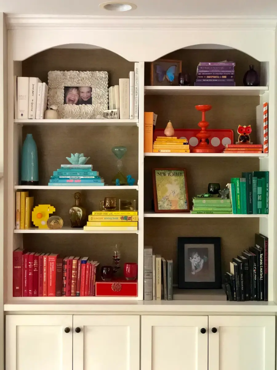 A bookshelf with books organized by color in rainbow order, decorative items including framed photos, small sculptures, a clock, and vases, all arranged neatly on white shelves above white cabinets.