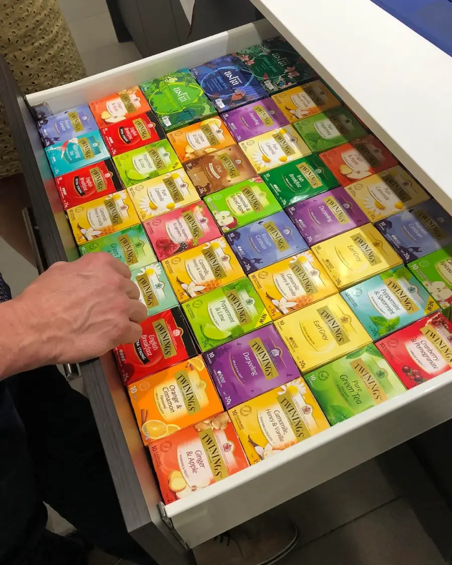 An open drawer neatly filled with rows of colorful Twinings tea boxes, organized by flavor and color. A person’s hand is reaching into the drawer to select a tea.