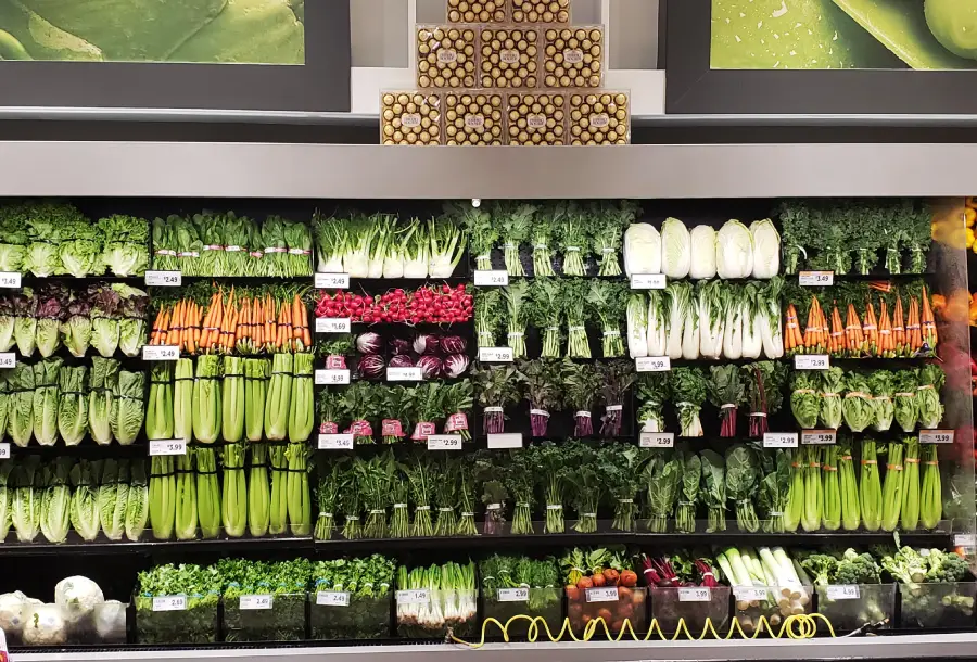 Colorful display of fresh vegetables arranged in neat rows at a grocery store, including celery, carrots, radishes, leafy greens, lettuce, and herbs, with price labels visible on the shelves.