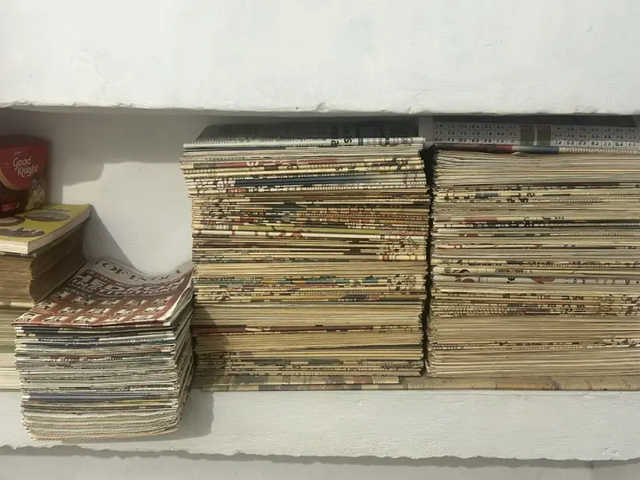 Stacks of old magazines or comic books are neatly piled on white shelves against a white wall, with a red cup visible on the left side of the image.