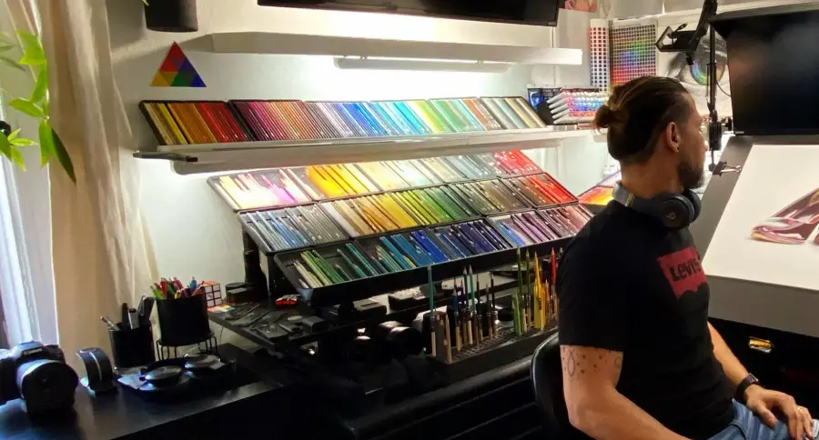A man with a ponytail and headphones around his neck sits at a desk filled with art supplies and neatly arranged colored pencils. The space is well-lit, organized, and decorated with a color wheel on the wall.