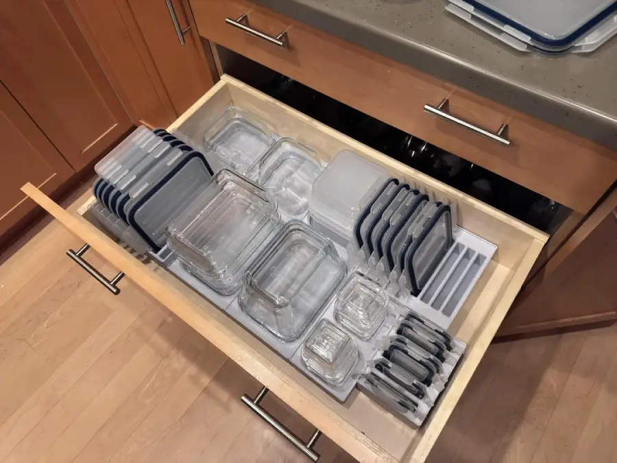 An open kitchen drawer neatly organized with glass food storage containers and matching lids, arranged by size. The containers are stacked vertically, with lids stored upright in dividers.