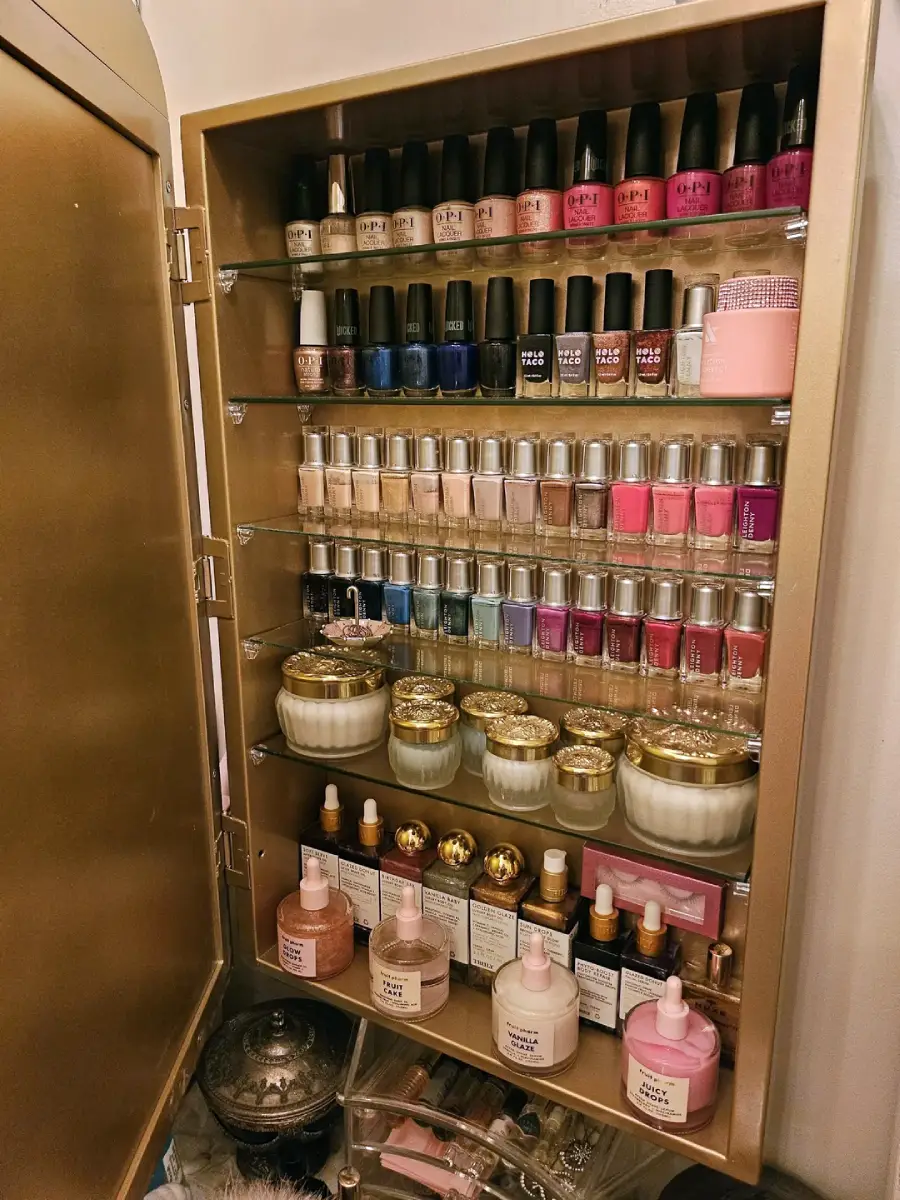 An organized gold cabinet with shelves holding rows of colorful nail polishes, skincare bottles, creams with gold lids, and beauty products arranged neatly inside.