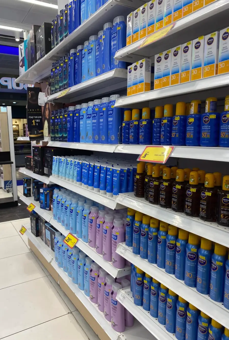 Shelves in a store filled with various brands and types of sunscreen lotions and sprays. Bright yellow sale tags are visible on some shelves, and the products are arranged neatly in rows by type and brand.