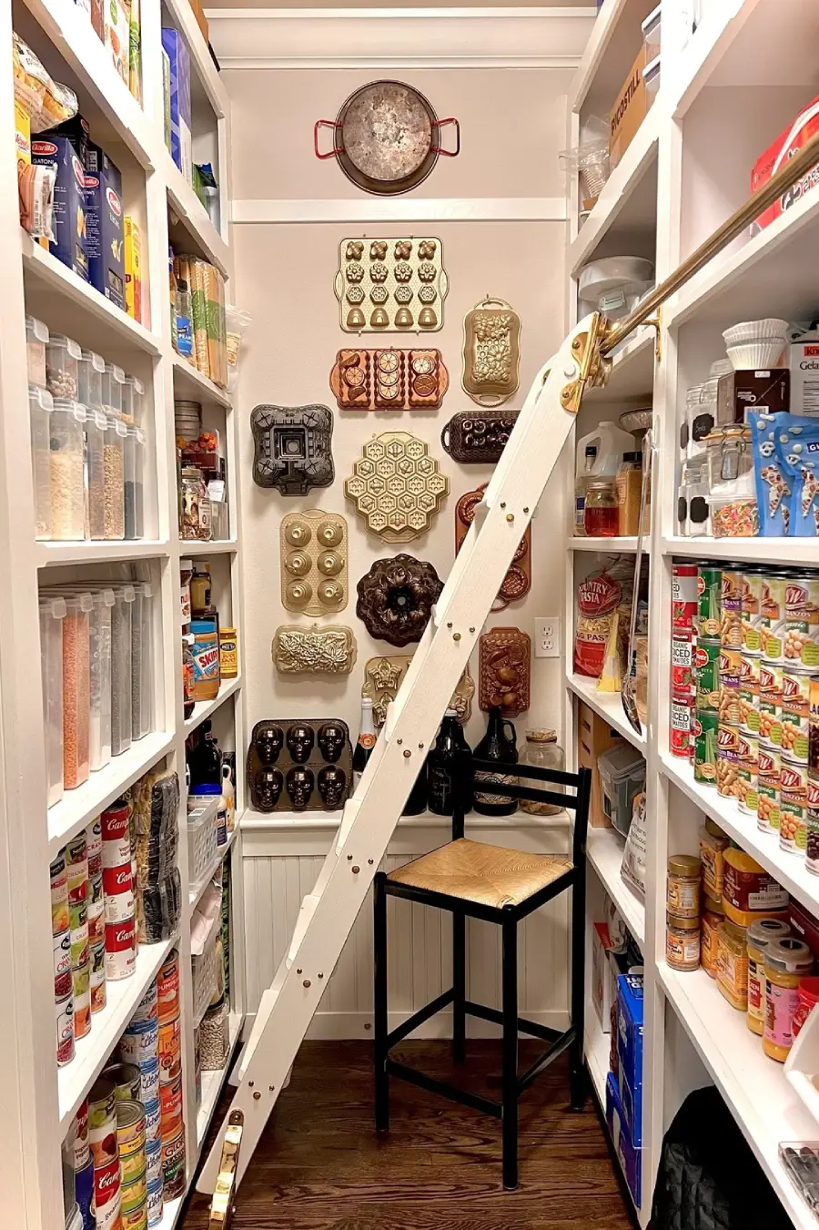 A well-organized pantry features labeled shelves of canned goods, snacks, and containers. A rolling ladder leans against the shelves, and vintage copper molds are displayed decoratively on the back wall above a chair.
