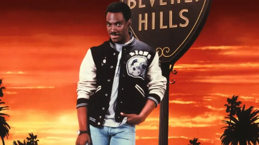 A man in a black and white varsity jacket and jeans stands in front of a Beverly Hills sign at sunset, with palm trees silhouetted in the background.