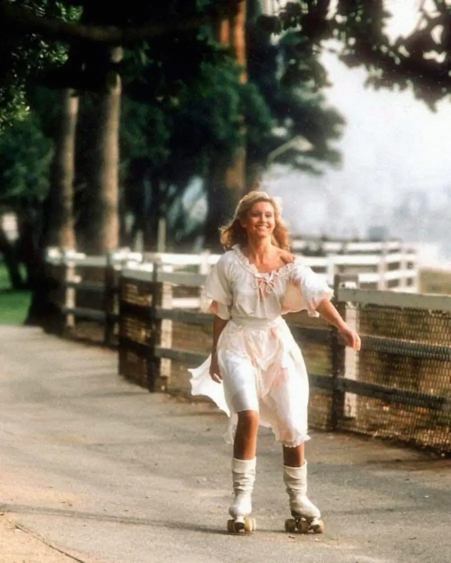 A woman in a white dress roller skates along a sunny, tree-lined path bordered by a wooden fence, smiling as she enjoys the outdoor scene.