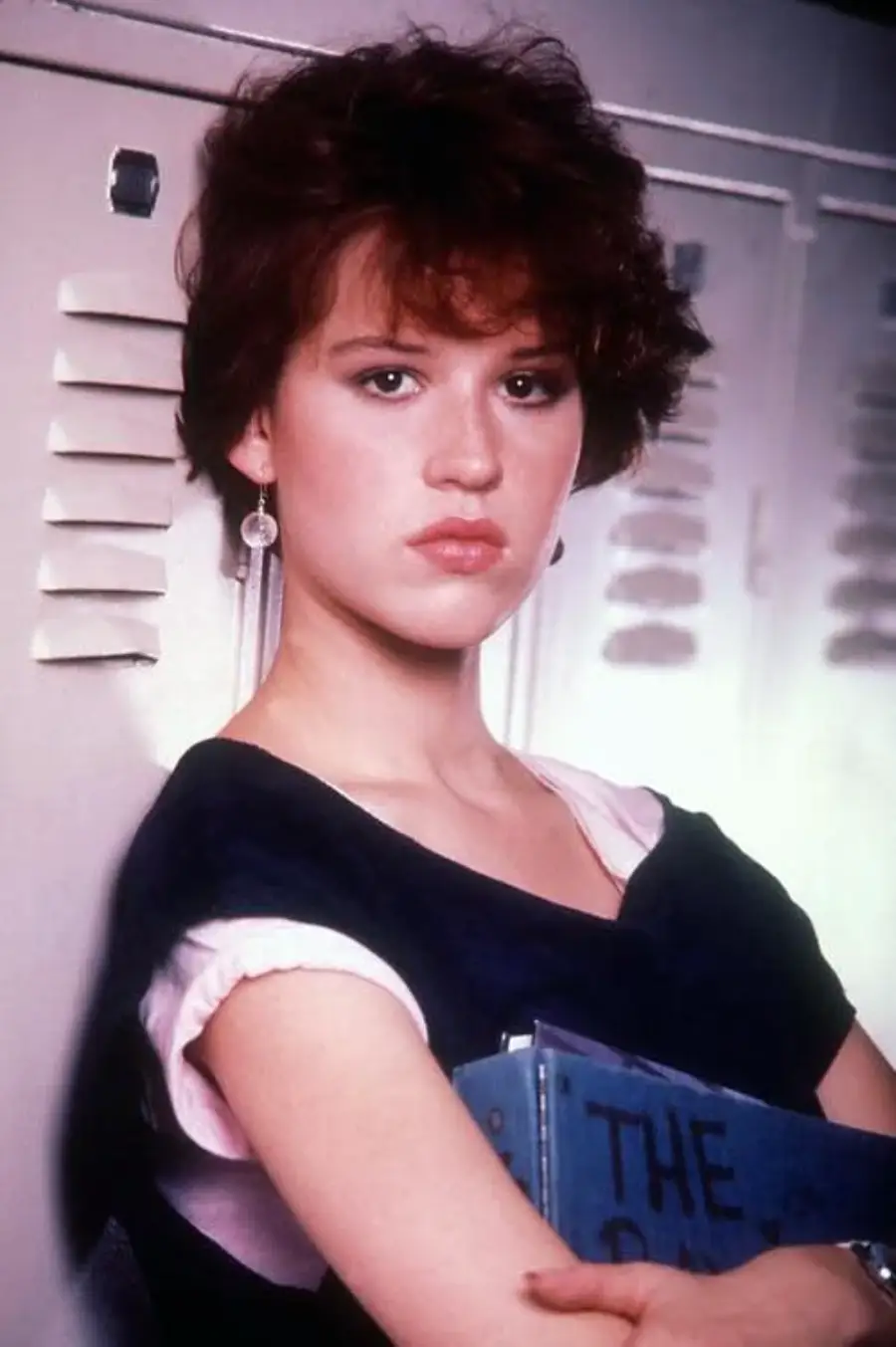 A young woman with short, curly brown hair stands in front of lockers, wearing earrings and a dark top layered over a light pink shirt, holding a blue book and looking seriously at the camera.