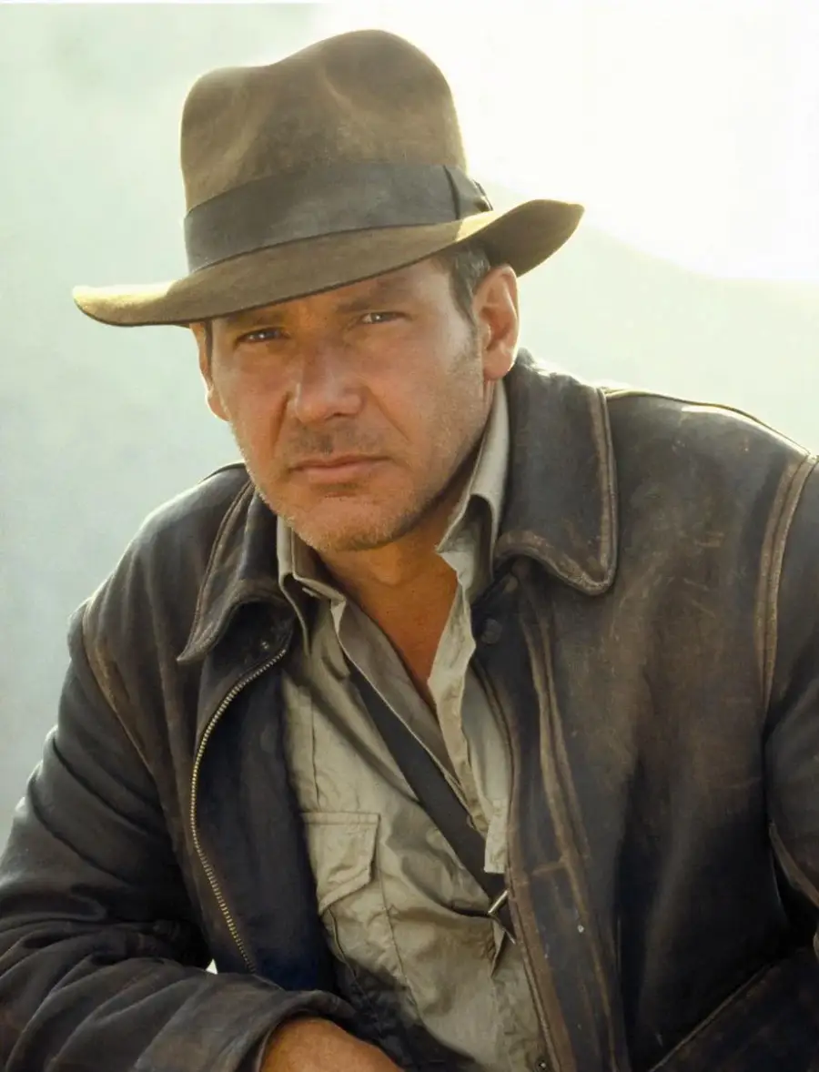 A man wearing a brown fedora hat, a rugged leather jacket, and a light shirt, looking intently at the camera against a light, blurred background.