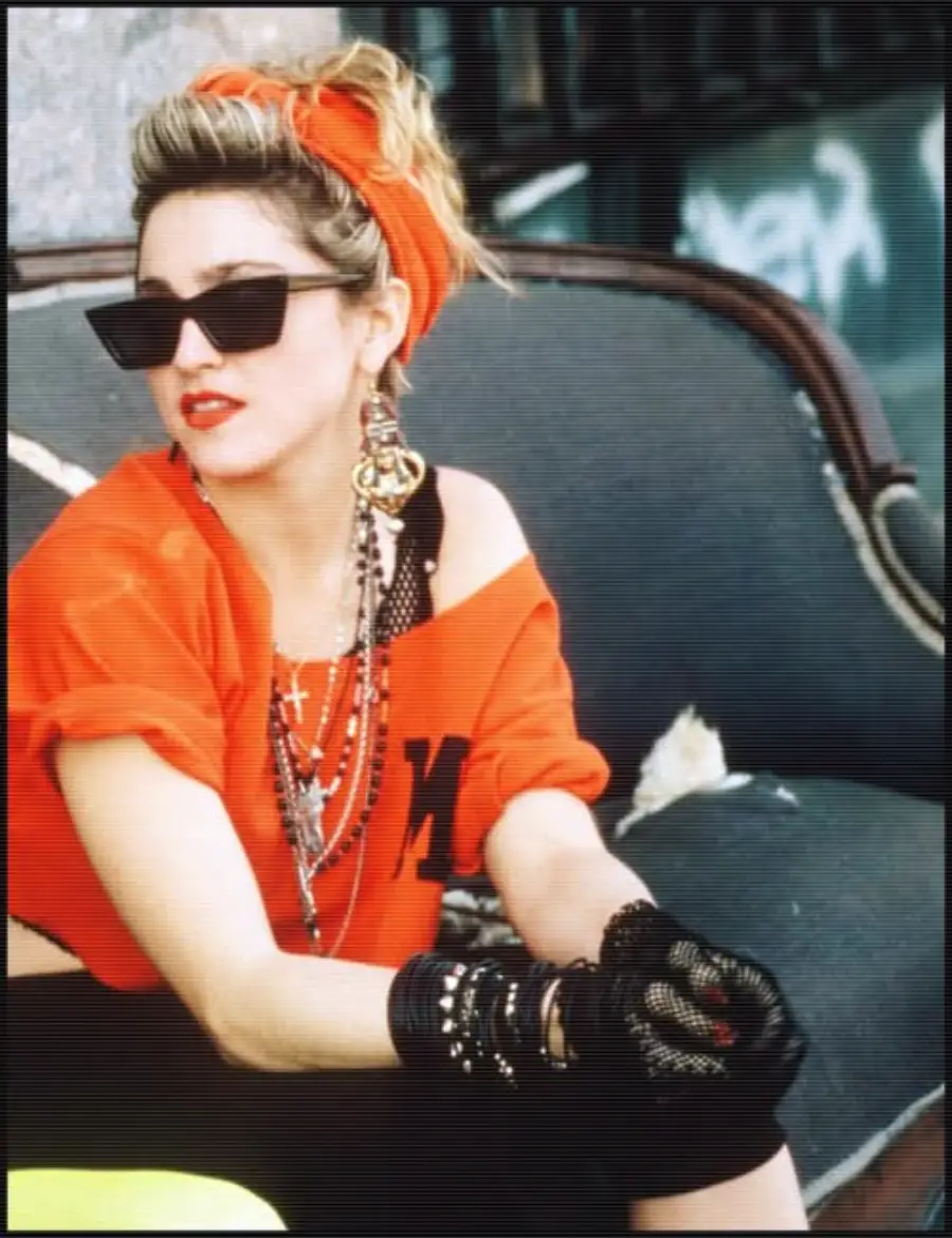 A woman in 1980s fashion sits on a worn sofa. She wears a red top, headband, black lace gloves, layered necklaces, large earrings, and dark sunglasses, with her hair styled up and red lipstick.