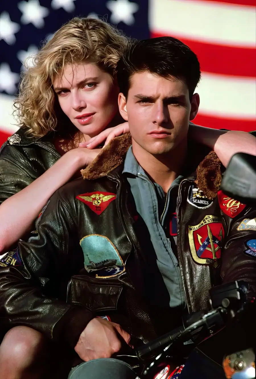 A man and woman in brown leather jackets pose together in front of a large American flag. The woman stands behind the man with her hands on his shoulders, while he sits in front, both looking directly at the camera.
