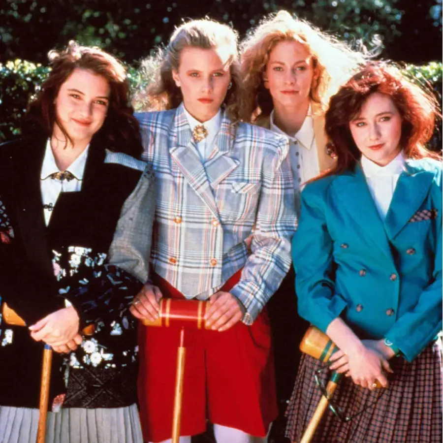 Four young women in colorful, bold 1980s fashion stand together outdoors, each holding a croquet mallet, with trees in the background. They appear confident and stylish.