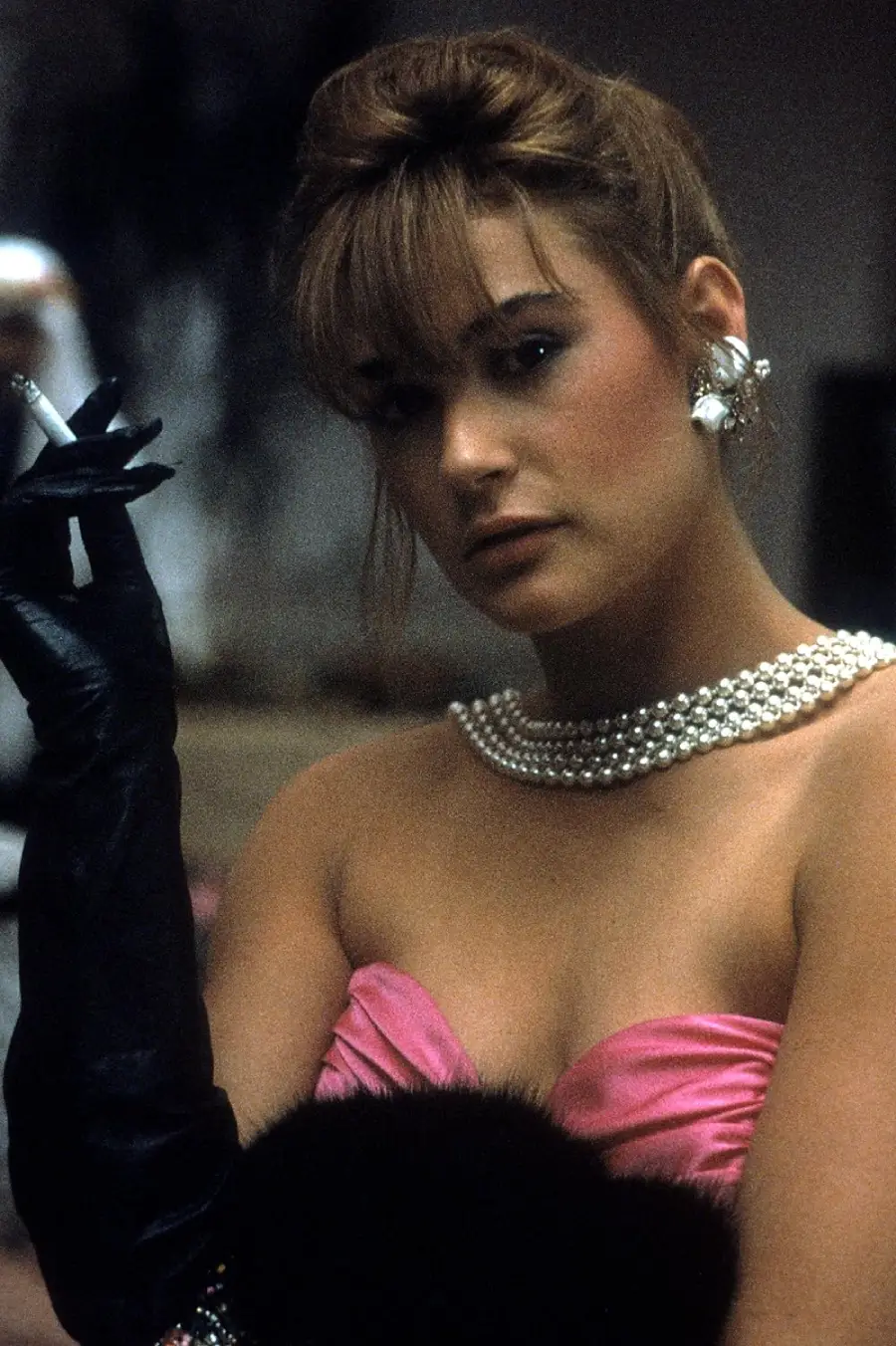 A woman wearing a strapless pink dress, layered pearl necklace, large earrings, and black evening gloves holds a cigarette. Her hair is styled in an updo with bangs, and she gazes toward the camera with a serious expression.