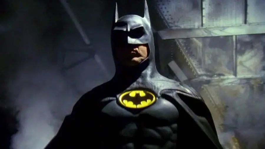 A person dressed as Batman stands in a dark, industrial setting with mist and dim lighting, wearing a black suit with a yellow bat symbol on the chest.