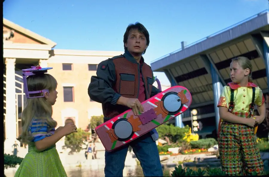 A young man in a futuristic outfit holds a pink hoverboard, standing outside with two girls dressed in colorful, unconventional clothing. Modern buildings and greenery are visible in the background.