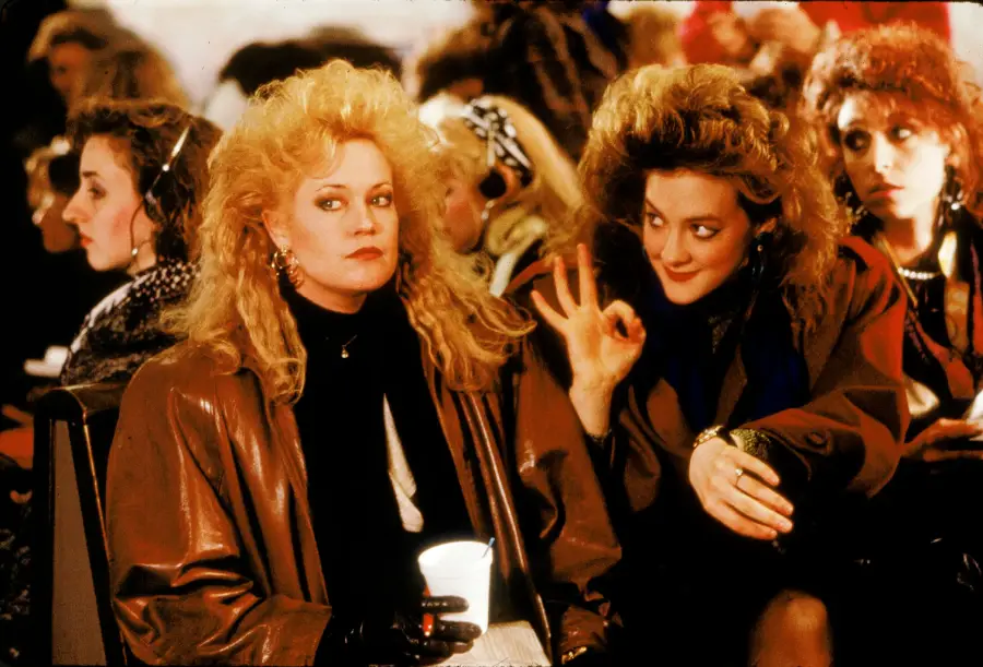 Two women with big, teased 1980s hairstyles sit close together. One, wearing a brown coat, stares forward holding a cup, while the other, also in brown, smiles and makes an "OK" hand gesture beside her face. Others sit in the background.