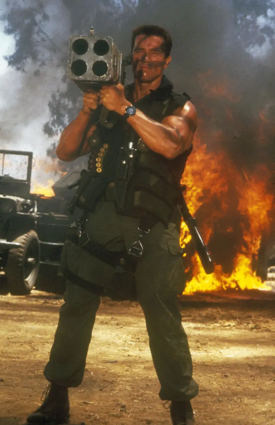 A muscular man in military gear stands in front of a burning vehicle, holding a large rocket launcher. He has a serious expression, black face paint, and wears a tactical vest with ammunition. Smoke and flames fill the background.