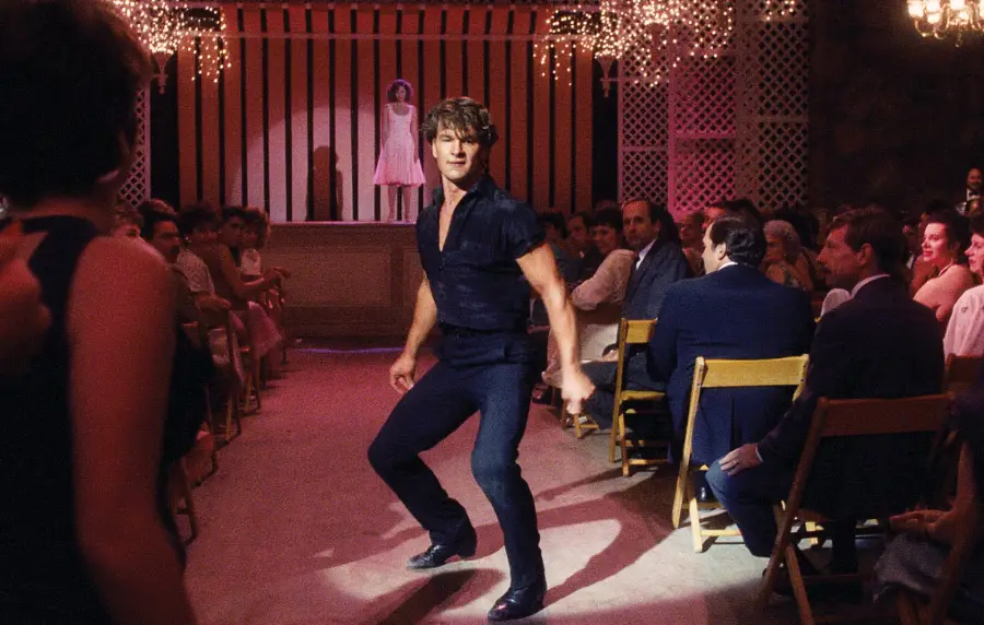 A man in dark clothing dances confidently in the center of a room lined with seated people, with soft lighting and chandeliers overhead, while a woman stands on stage in the background.