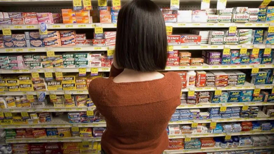 A person with shoulder-length dark hair, wearing a rust-colored sweater, stands in a pharmacy aisle facing shelves stocked with various over-the-counter medicines and pain relievers.