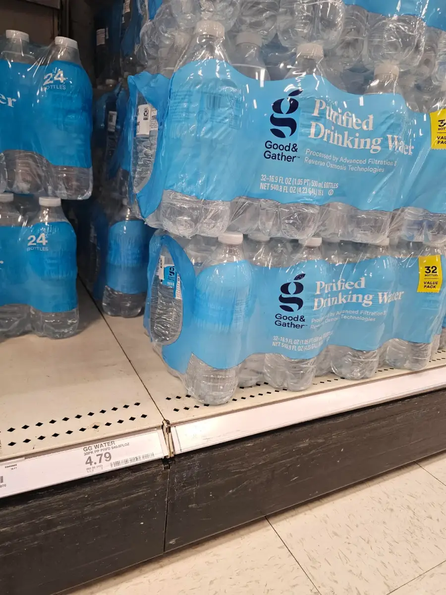 Cases of Good & Gather purified drinking water bottles stacked on a store shelf, with a price tag of $4.79 visible below them. The blue packaging wraps around 24-bottle packs.
