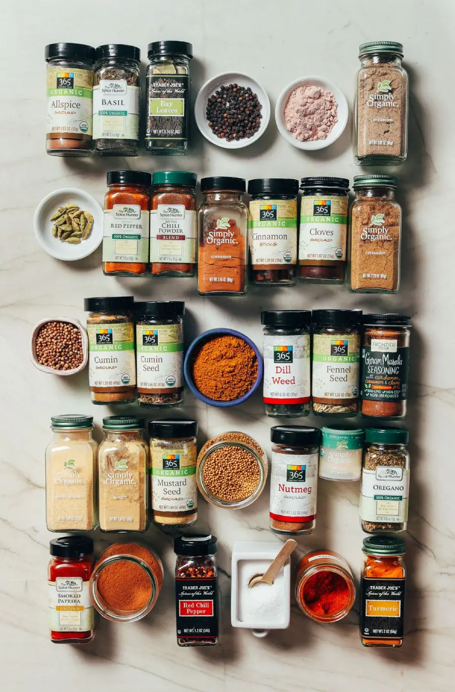 A variety of spice jars and containers are neatly arranged on a white marble surface. The collection includes herbs, ground spices, seeds, and small bowls of spice blends, all labeled and organized in rows.