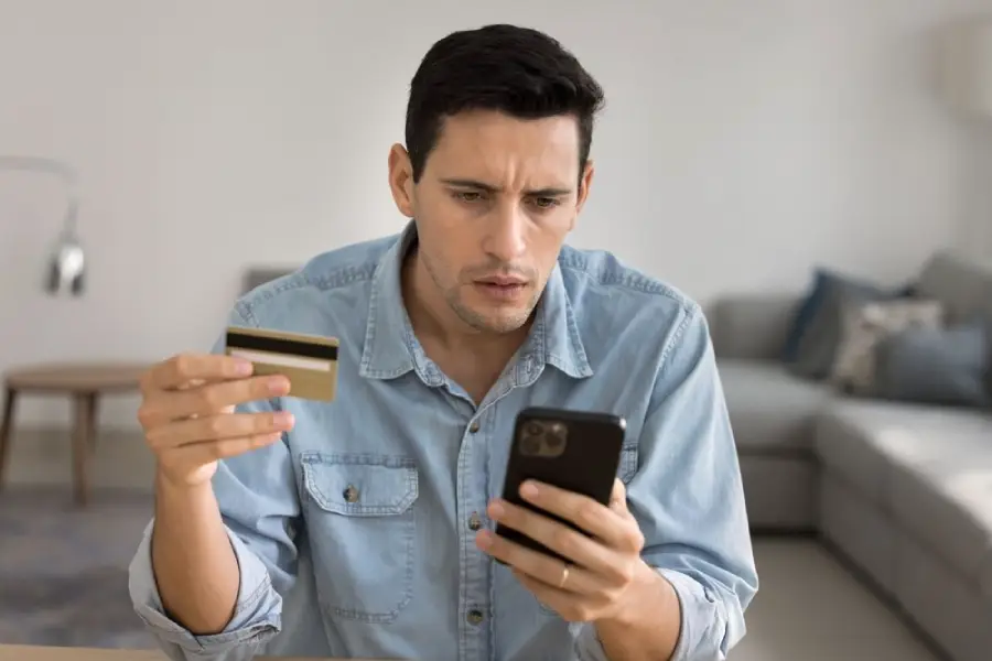 A man in a denim shirt sits indoors, holding a credit card in one hand and looking concerned at his smartphone with the other hand. A sofa and lamp are visible in the background.
