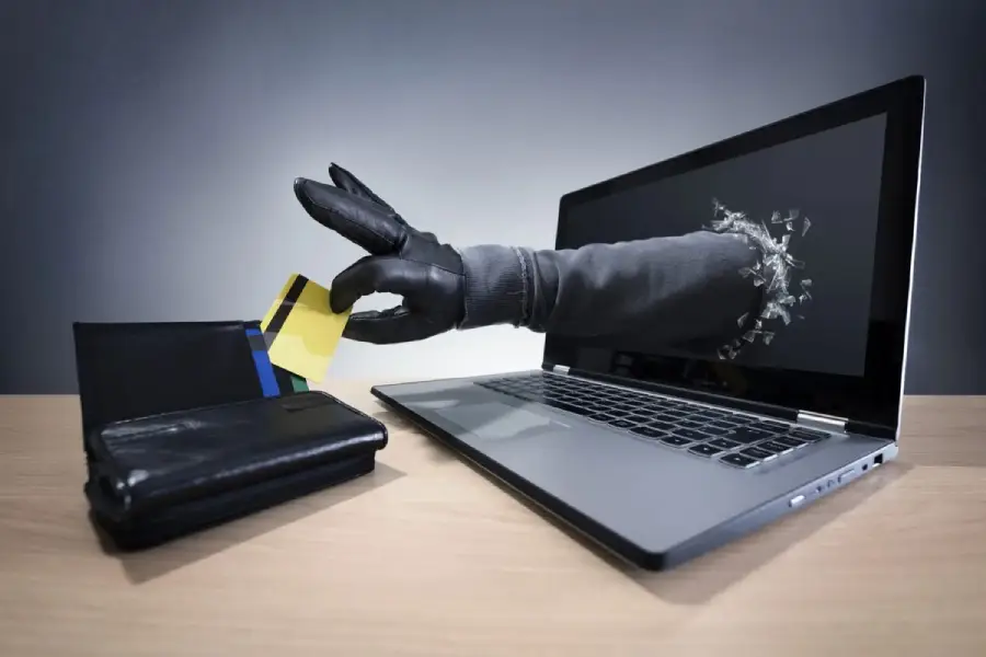 A gloved hand emerges from a laptop screen, reaching for a credit card next to a wallet on a desk, symbolizing online theft or cybercrime.