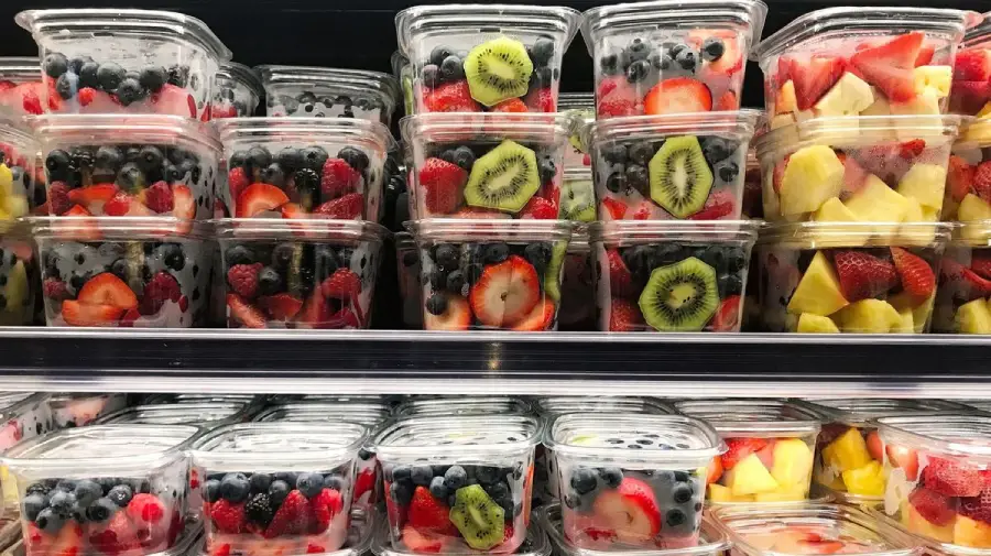 Plastic containers filled with assorted fresh fruit, including strawberries, blueberries, kiwi slices, and pineapple, stacked neatly on refrigerated grocery store shelves.