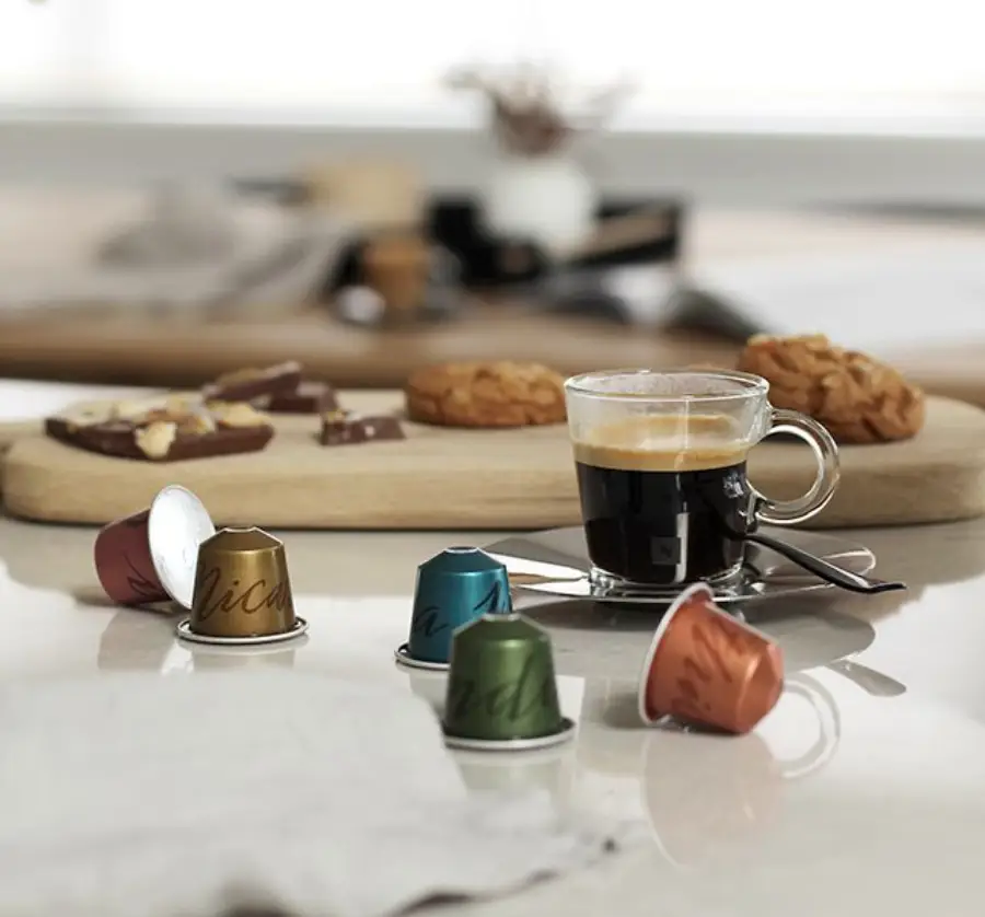 A glass cup of espresso sits on a saucer with a spoon, next to colorful coffee capsules. In the background, pastries and chocolate pieces are arranged on a wooden board.