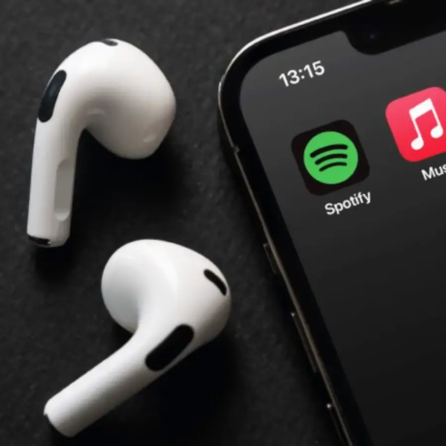 A pair of wireless white earbuds lie next to a smartphone displaying the Spotify and Music app icons on its screen. The time on the phone shows 13:15. The background is a dark surface.