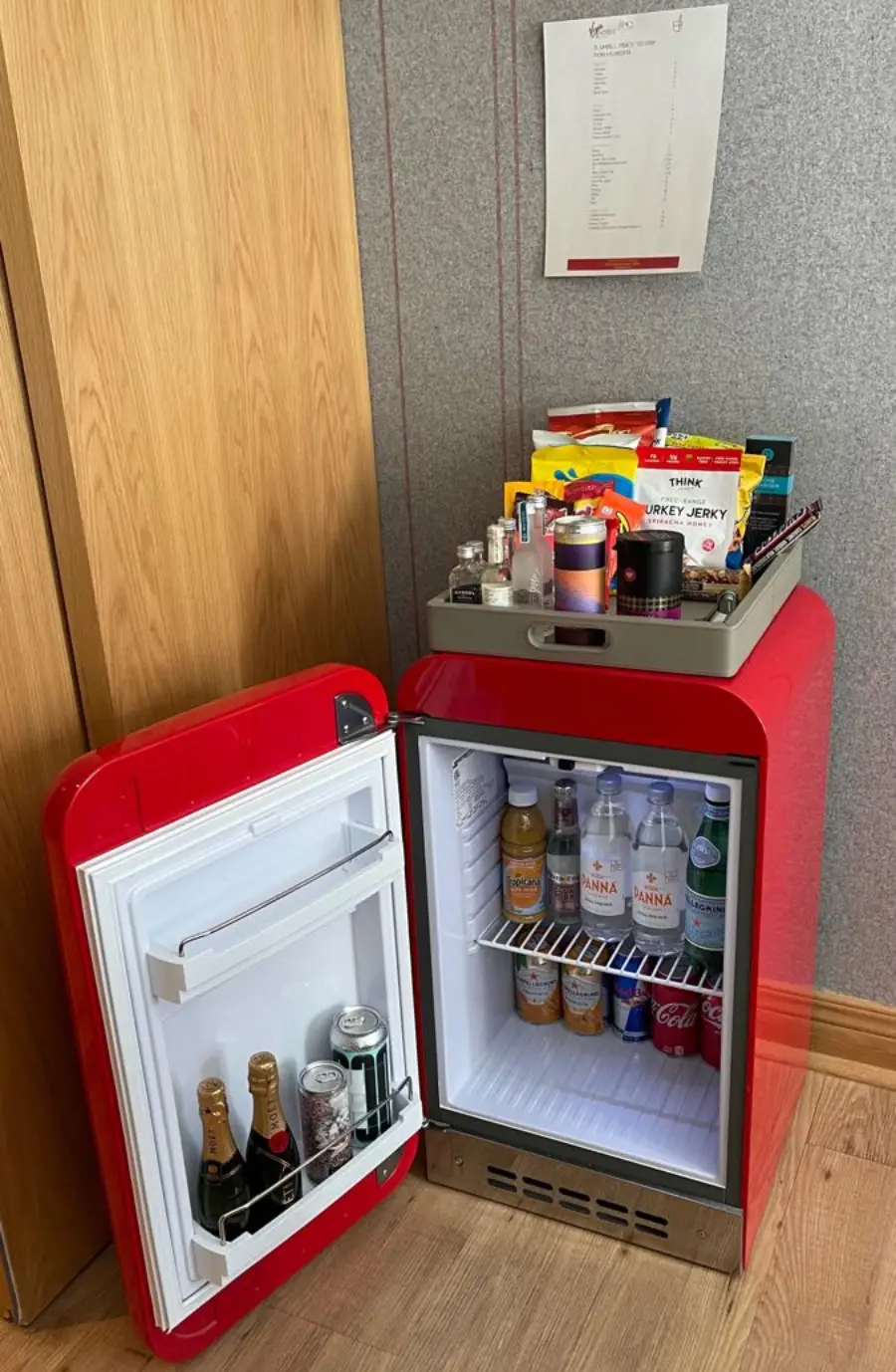 A red mini fridge with its door open, showing bottled drinks, soda cans, and water inside. On top of the fridge is a tray with snacks and more drinks, and a document is taped to the wall above.