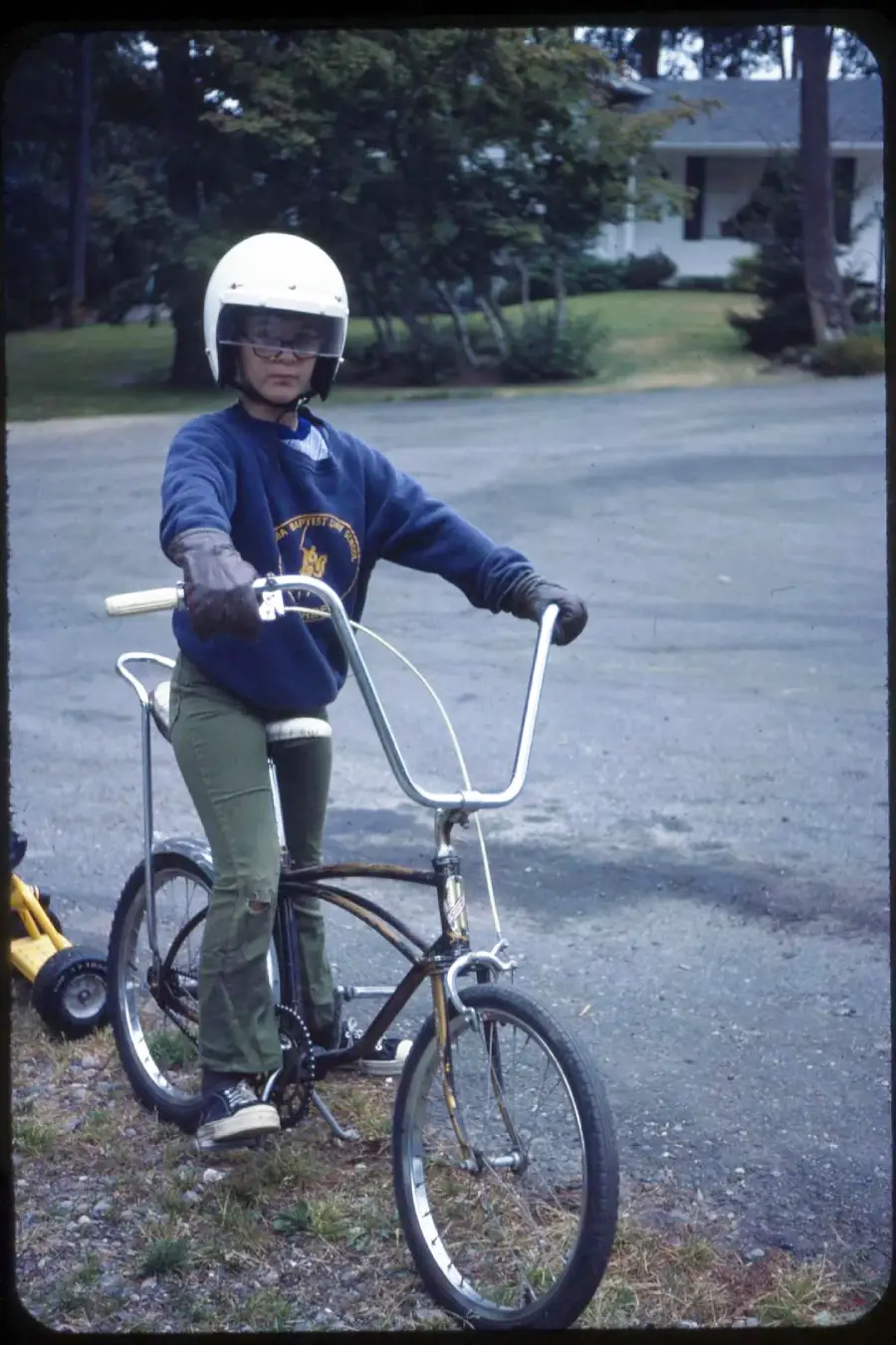 A person wearing a white helmet, glasses, gloves, and a blue sweatshirt stands on a vintage bicycle with high handlebars in a driveway, with a yellow toy vehicle and a house in the background.