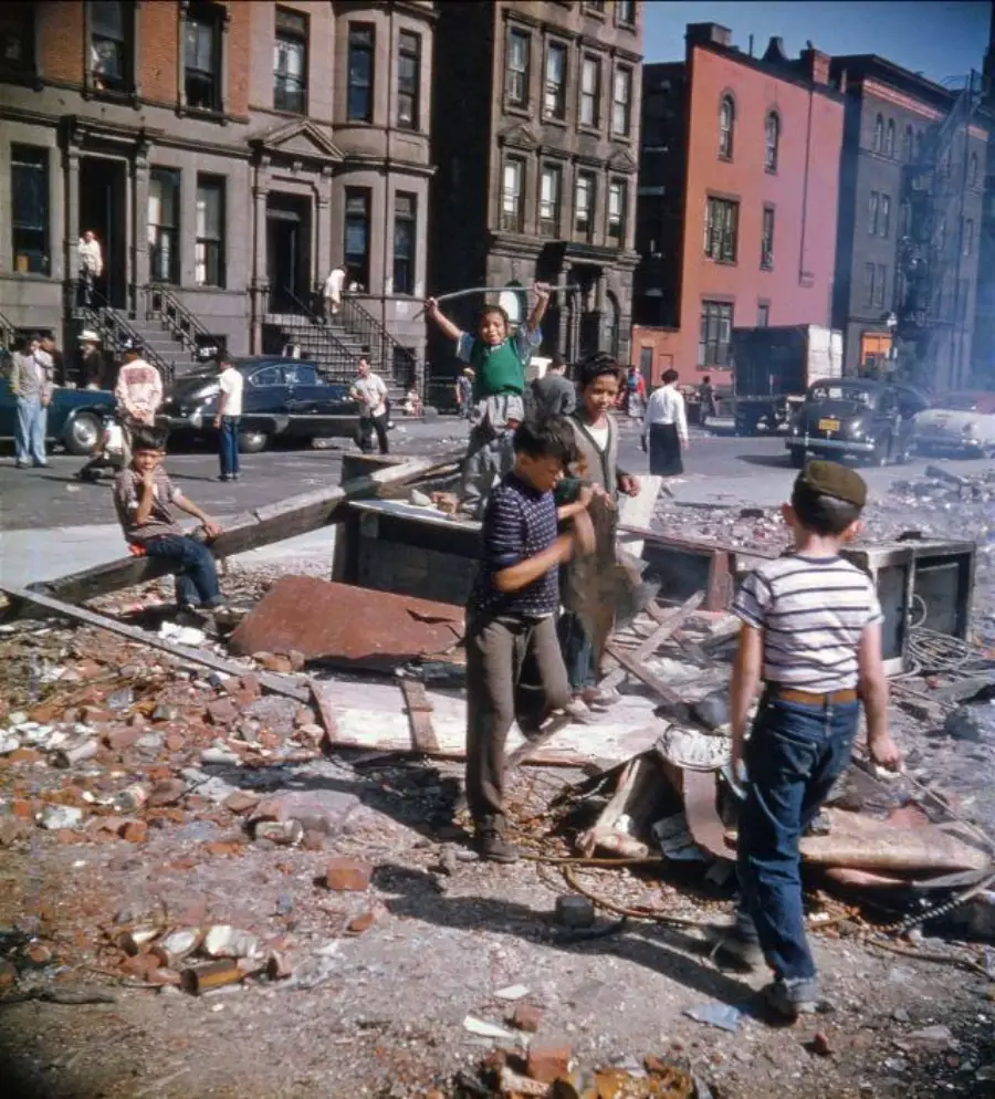 Children play among rubble and debris on a city street, with brownstone buildings and parked cars in the background; several people watch from stoops and sidewalks on a sunny day.