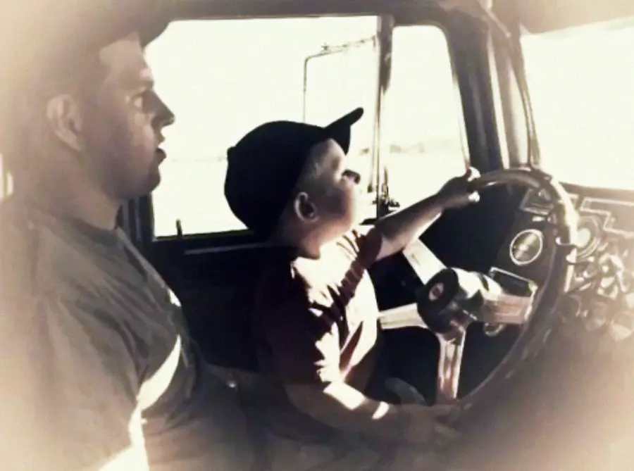 A sepia-toned photo shows a man and a young boy sitting together in a vehicle. The boy, wearing a cap, is holding the steering wheel while the man sits beside him, watching.
