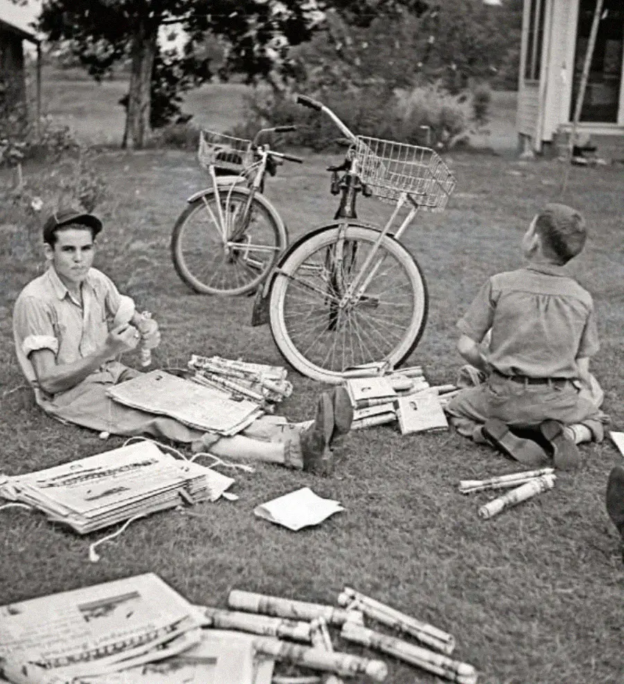 Two boys sit on the grass surrounded by rolled and flat newspapers, preparing them for delivery. Two bicycles with baskets are nearby, and a house and trees are visible in the background.