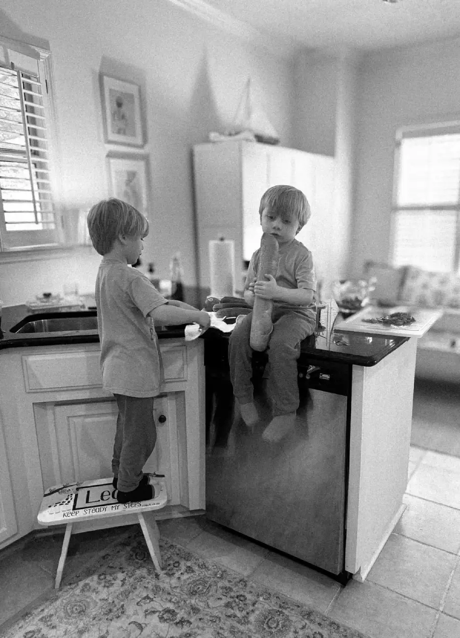 Two young children, one standing on a step stool by the kitchen sink washing dishes, and the other sitting on the countertop holding a baguette. The kitchen is bright and homey with natural light coming in.
