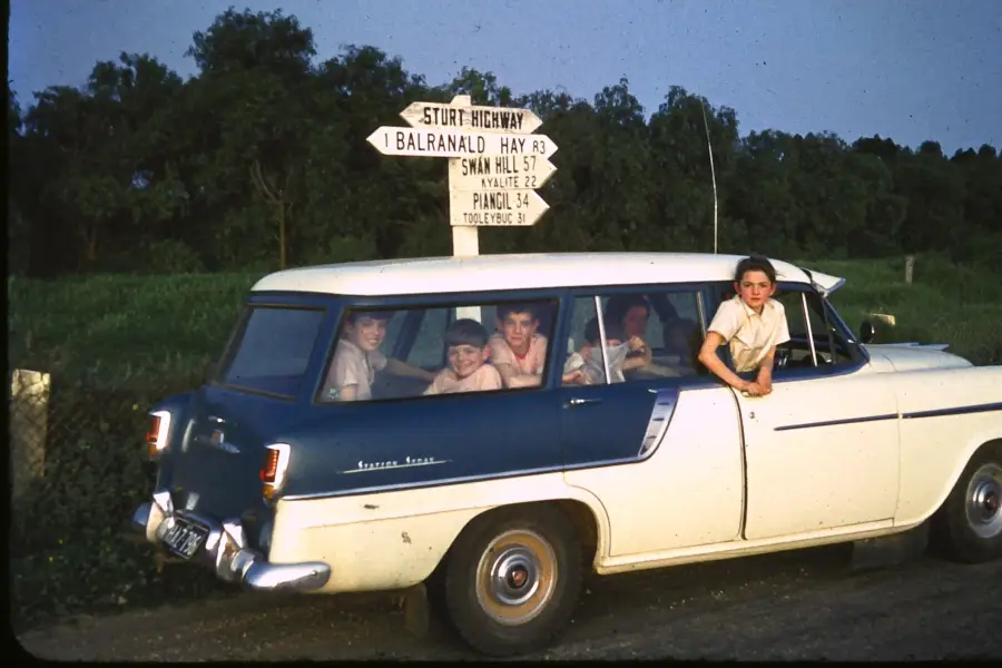 A group of children sit inside a vintage blue and white station wagon parked beside a road, with one boy leaning out the window. Road signs behind the car point toward various towns on the Sturt Highway.