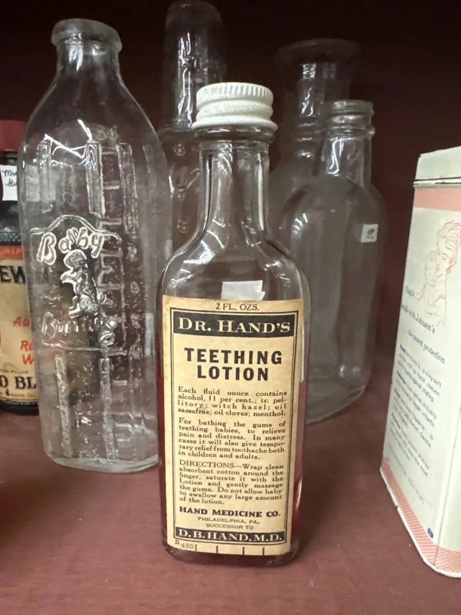 A vintage glass bottle labeled "Dr. Hand's Teething Lotion" sits on a shelf among other old bottles and containers. The label details its use for soothing teething pain in babies.