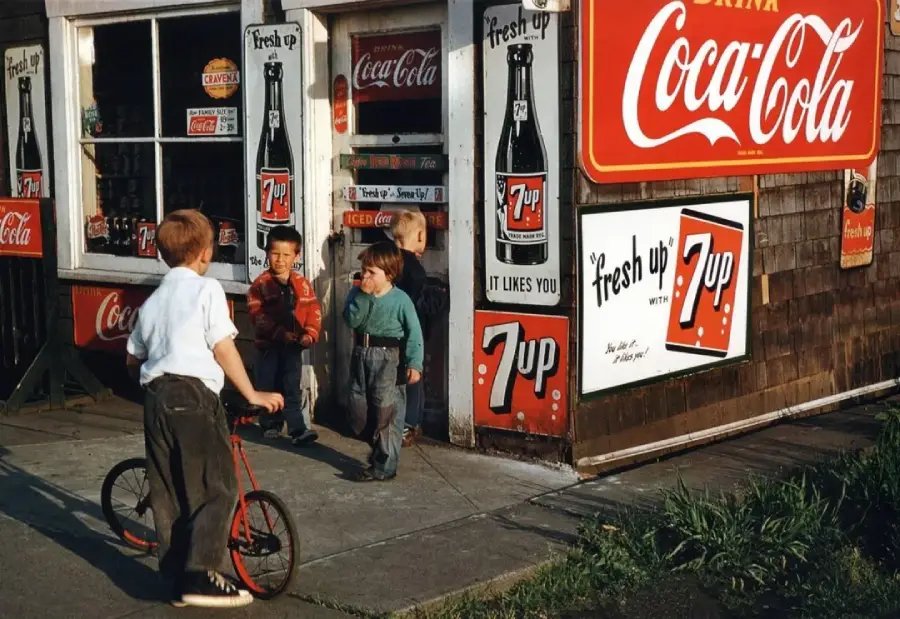 Four young boys, one with a red scooter, talk and laugh outside a vintage corner store covered in Coca-Cola and 7 Up signs. The scene appears nostalgic, evoking a small-town, mid-20th century atmosphere.
