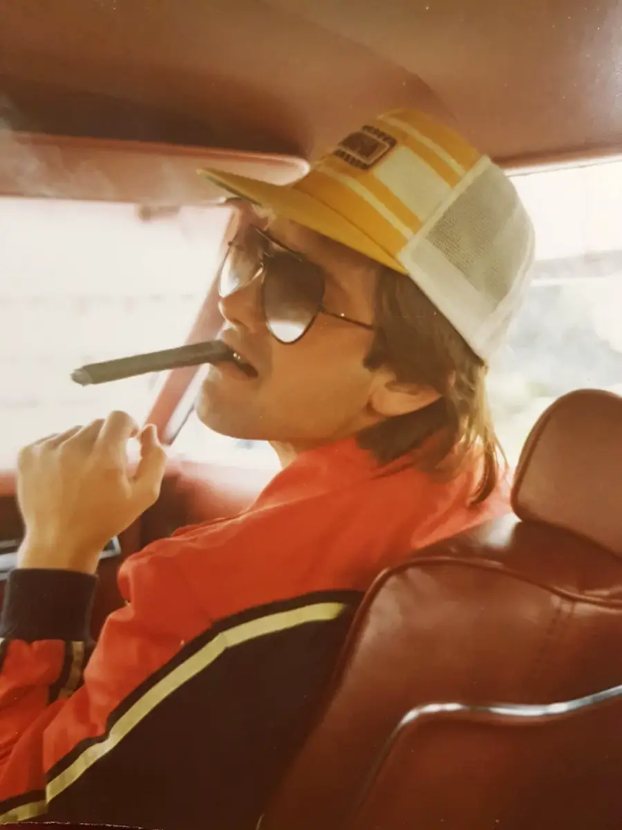 A person in a red jacket and yellow cap sits in a car, wearing large sunglasses and holding a cigar in their mouth, looking toward the camera.