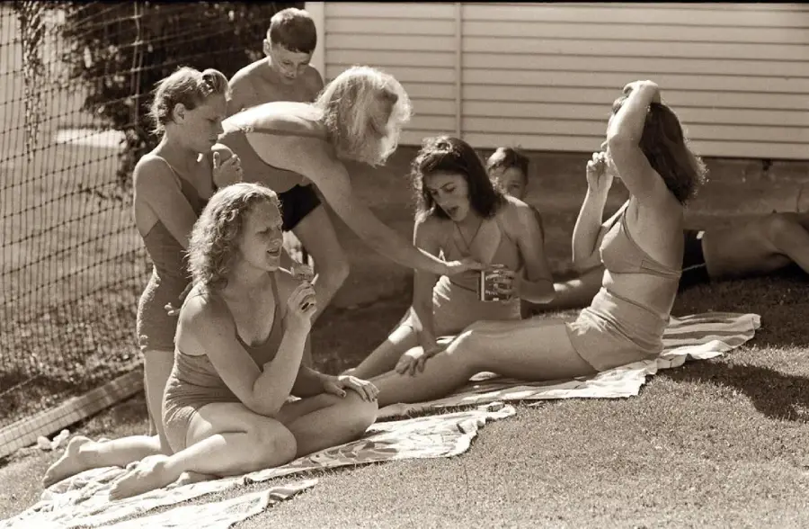 A group of women and children in swimsuits relax on towels outdoors, enjoying a sunny day. One woman applies lotion to another, while others sit and chat on the grass beside a building.