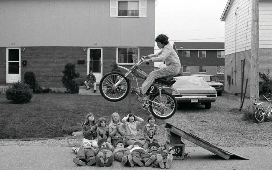 A boy on a bicycle jumps off a makeshift ramp over a group of kids lying on the ground, while other children watch in a suburban neighborhood.